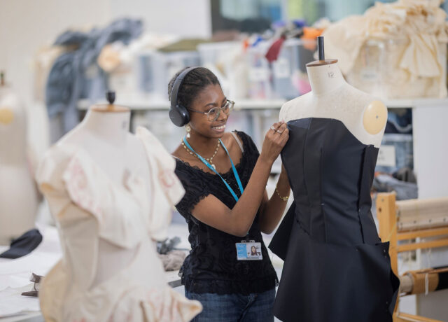 Student sewing a dress on a mannequin in a class room surrounded by garments, fabrics and equipment