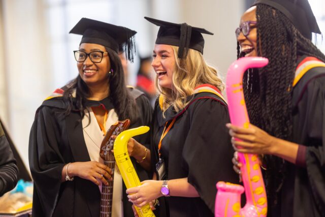 3 students in graduation dress smiling and holding inflatable musical instruments