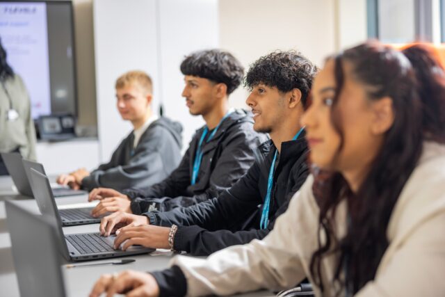 Three male and one female student sitting at a desk working on laptops