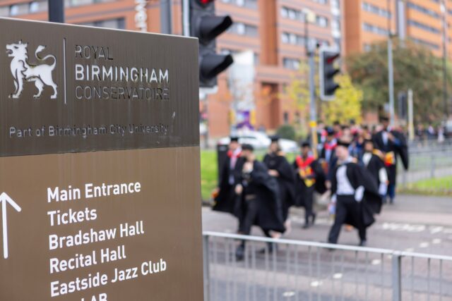 Royal Birmingham Conservatoire signage with out of focus background of students in graduation dress in crossing a road.
