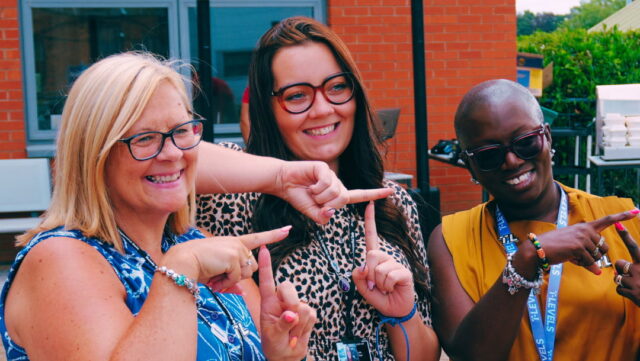3 members of staff making T signs with their fingers, standing outside with a building the the background