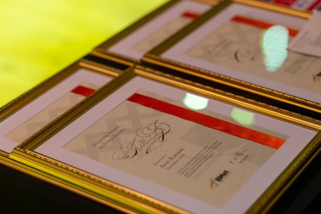 Gold framed award certificates stacked on a black surfaced table