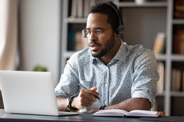 Man with headphones and floral shirt sitting at desk with notebook and holding pen and looking a laptop