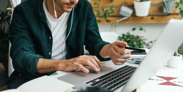 Man with earphones and with green shirt sitting at desk working on a laptop and holding pen