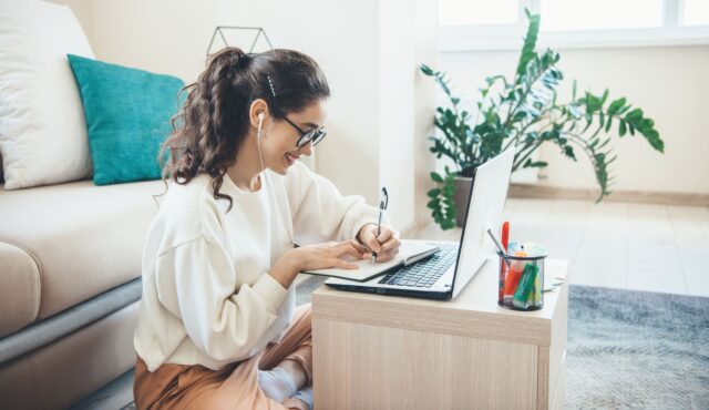 Female sitting on floor with earphones looking at laptop and making notes with pen and notebook, in a furnished domestic room