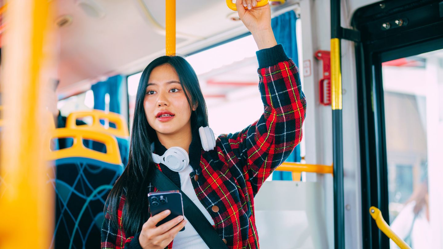 Female with checked coat and headphones standing on a bus holding handrail and holding mobile phone
