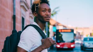 Male with white T-shirt and grey backpack standing in street with red bus in background
