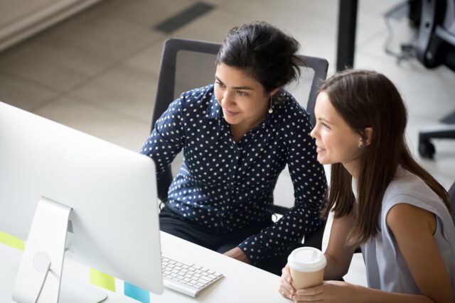 Two females sitting at a desk looking at a computer screen, one is holding a drinks cup