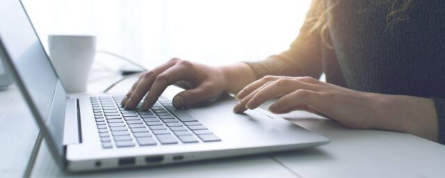Female sitting at a desk typing and moving the mouse pad on a laptop in a room with a white background
