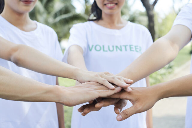 Five arms held out with their hands stacked together. The people are wearing white t-shirts with the word VOLUNTEER printed in green on them