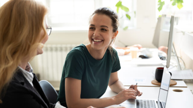 Female in green t-shirts sitting at a desk with laptop and holding pen smiling at another female sitting beside her