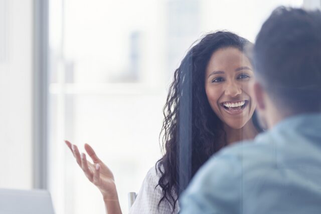 Female smiling and talking to at another person who has their back to the camera, in a bright room