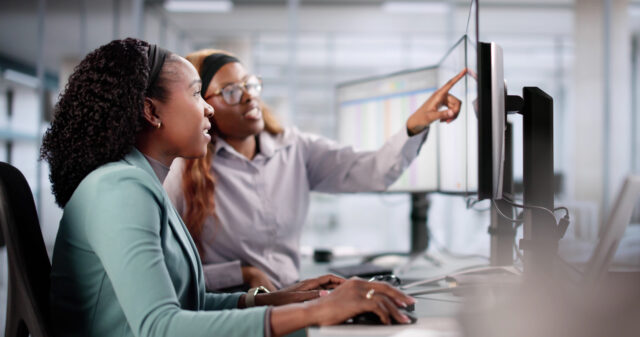 Two females sitting at computer with three screens. One female is pointing to something on the right screen