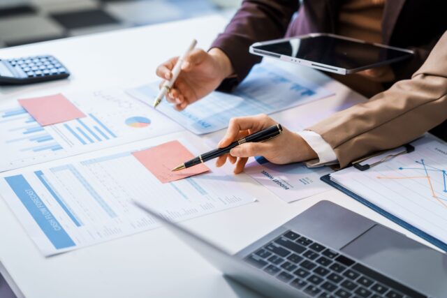 Close up of hands of two people holding pens reviewing printed charts and graphs at a desk, using a tablet, laptop, and calculator.