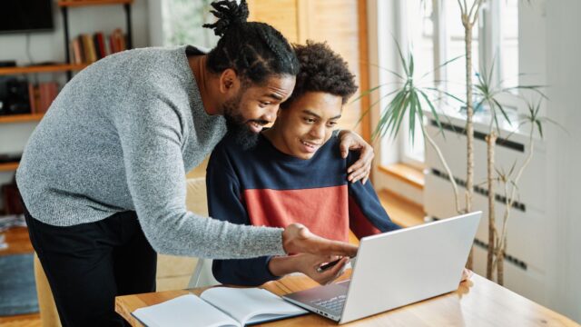 A smiling man talks to a seated young person working at a laptop, pointing to the screen in a bright room